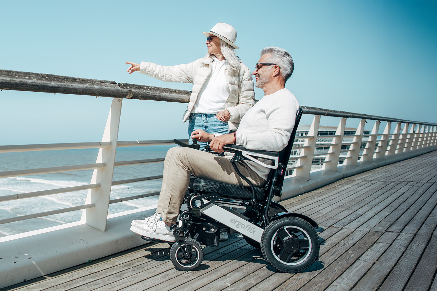 Un señor mayor sentado al sol en su silla de ruedas ergoflix® LX contempla el mar junto a su pareja.